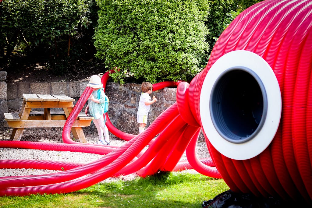 two children speaking inot the tentacles of a giant red squid sculpture in the PK Porthcurno Gardens.